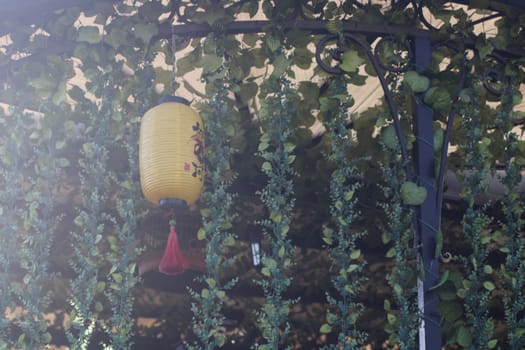 A bright yellow lantern dangles gracefully among lush green vines in a tranquil outdoor setting, bathed in afternoon sunlight, creating a peaceful atmosphere.
