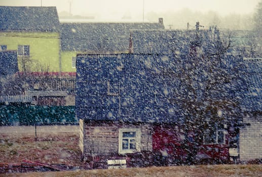 The image shows a small village on a gloomy winter day. The snow is falling heavily, and the houses are covered in snow. The image has a cold and desolate feel to it.
