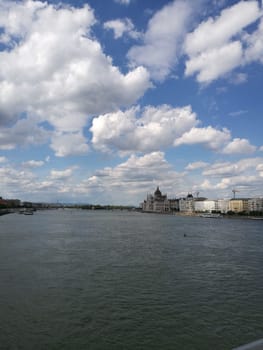 A tranquil river flows gently beneath a sky filled with fluffy clouds, while charming buildings line the water's edge in a vibrant cityscape.