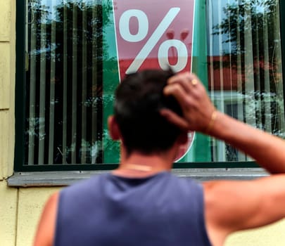 A man is standing outside a store, scratching his head as he observes a large percentage sign displayed in the window. His posture suggests he might be puzzled or considering the implications of the advertised discount, sale, or promotion.