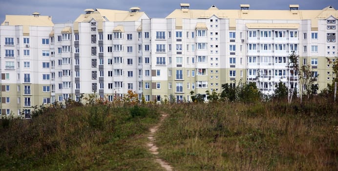A tranquil path meanders through a lush green area, leading directly to a cluster of modern apartment buildings that dominate the landscape. The buildings display a mixture of soft pastel colors and large windows, reflecting contemporary architectural styles. The sky overhead is filled with moody clouds, hinting at an impending change in weather.