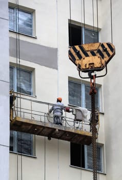 A construction crew diligently attends to the facade of a multi-story building, showcasing their expertise in exterior renovations. Suspended on a scaffold, one worker is meticulously applying finishing touches while another operates a crane above, ready to deliver necessary supplies.