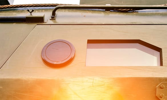 A close-up of a military vehicle featuring a square window and a round hatch, illuminated by sunlight, highlighting its robust exterior.