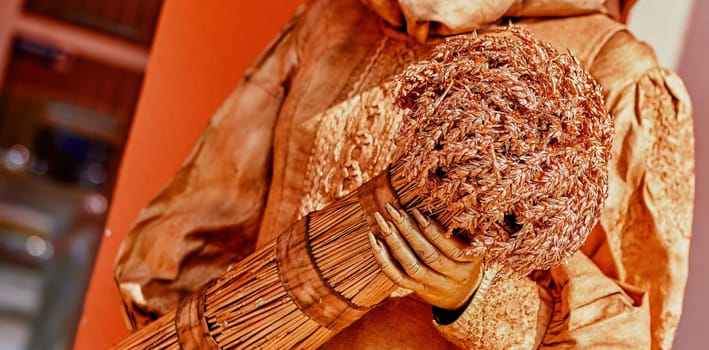 A captivating figure, adorned in textured materials, clutches a bundle of dried flowers, set against an orange backdrop in a busy cultural exhibition.