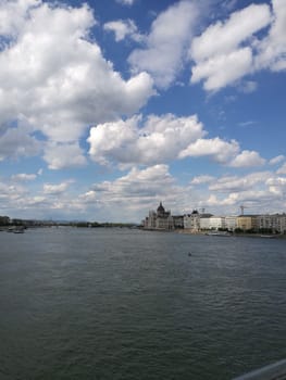 A stunning view captures the Danube River flowing serenely, framed by fluffy white clouds and an expansive skyline of Budapest as the sun begins to set.