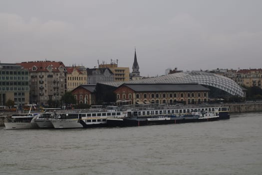 Several cruise ships are tied up along the riverbank, alongside a city filled with historic structures and a modern architectural dome under a cloudy sky.