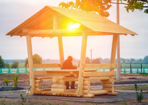 A lone individual relaxes under a wooden gazebo as the sun sets, creating a tranquil atmosphere in a rural park setting.
