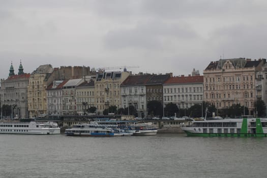 Several boats are moored along the river in Budapest, surrounded by beautiful historical architecture under an overcast sky.