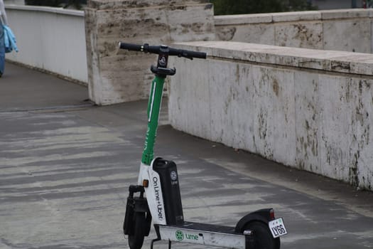 An electric scooter stands unused on a city sidewalk, showcasing a moment in urban mobility as pedestrians pass by in the background, highlighting modern transportation.