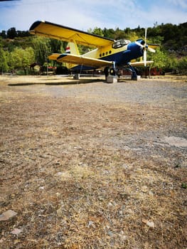 A bright yellow and blue vintage aircraft rests on gravel in a rustic area, framed by trees under a clear sky. The inviting atmosphere hints at historic aviation.