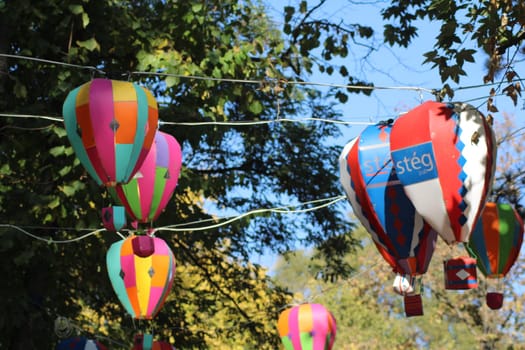 Brightly colored hot air balloons dangle between trees at a lively outdoor festival in early autumn, adding an enchanting touch to the atmosphere and delighting visitors.