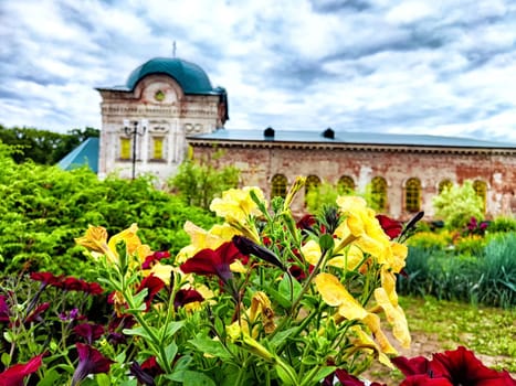 Colorful flowers thrive in a vibrant garden with an old building in the background, set against a cloudy sky.