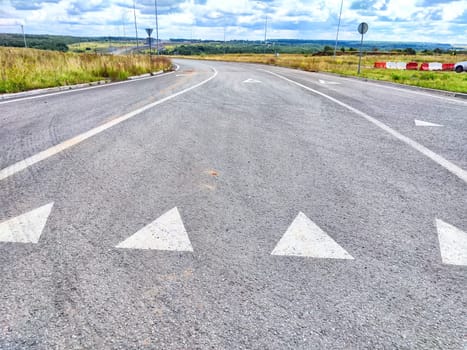 A winding road with visible arrows directing traffic, surrounded by open fields and a cloudy sky, indicating a rural setting.