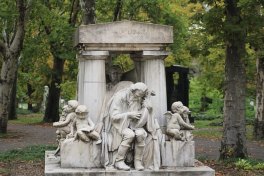 In a peaceful park, a beautifully carved marble monument features a grieving figure surrounded by cherubs, with vibrant autumn foliage in the background.