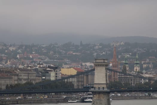 On a cloudy day, the Chain Bridge connects Buda and Pest, revealing views of Budapest’s distinct architectural styles and the flowing Danube River in the background.