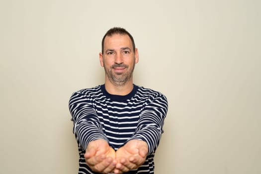 Portrait of a handsome, friendly bearded man offering something in his hands with arms outstretched. Studio shot isolated on beige background.