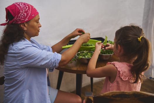 A mother and daughter sit together at a table, preparing a fresh leafy salad. The scene captures a warm family moment emphasizing healthy cooking and teamwork in a cozy home kitchen.