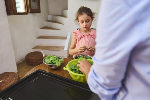 A young girl is seen assisting in the kitchen, preparing fresh greens with focus and care. The scene conveys a sense of family bonding and healthy living in a cozy home setting.
