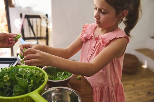 A young girl in a pink dress prepares a fresh salad in a cozy kitchen. Her focused expression and careful hands convey the joy of learning and healthy living in a family environment.