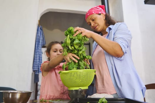 A mother and daughter wash fresh spinach in the kitchen, sharing a special bonding moment. The vibrant greenery and warm setting emphasize family, nutrition, and togetherness.