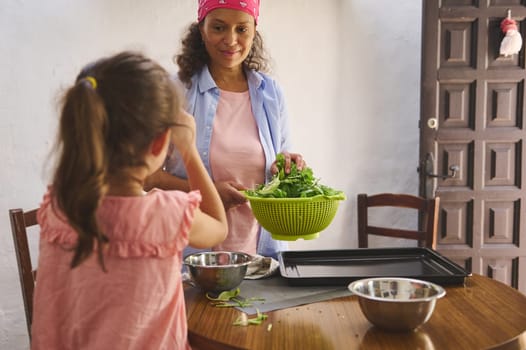 Mother and daughter enjoying quality time in the kitchen preparing a fresh green salad. The image captures a warm, family moment with a focus on health and togetherness.