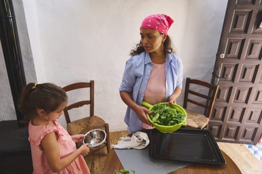 A mother and daughter spend quality time together in the kitchen, preparing a fresh green salad. The mother holds a colander filled with leafy greens while the daughter helps with a bowl.