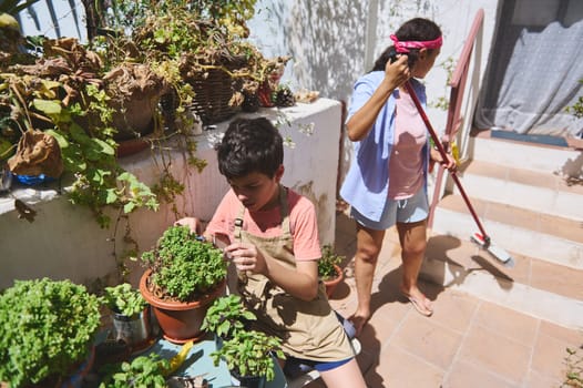 A young boy carefully tends to potted plants on a sunny patio while a woman sweeps nearby, creating a serene outdoor atmosphere.