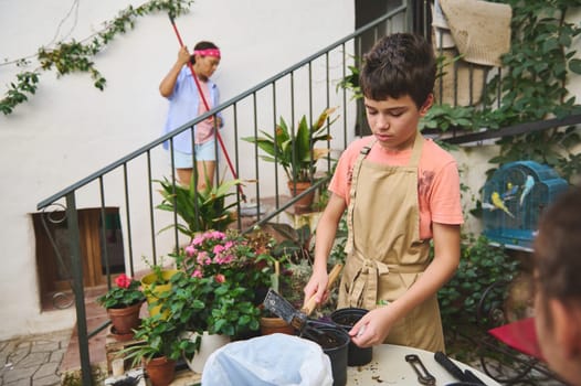 A young boy is diligently gardening with various tools, blending with a lush green garden. In the background, a woman is sweeping an outdoor area, symbolizing teamwork and family bonding.