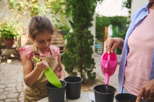 A young girl and an adult engage in gardening activities outdoors. The child focuses on planting while the adult waters a pot, creating a nurturing and educational moment in nature.