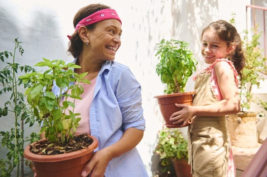 A joyful mother and daughter bond while gardening, holding potted plants in a sunlit garden, sharing smiles and happiness.