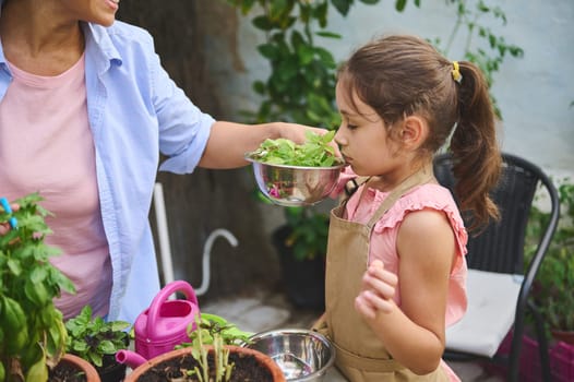 A young girl in an apron enjoys smelling fresh herbs while gardening with family. The setting is a lush, green backyard, emphasizing nature's beauty and family bonding through hands-on learning.