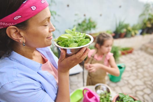 A mother appreciates the aroma of fresh basil from the garden while her daughter assists nearby, embodying a sense of family, nature, and togetherness.