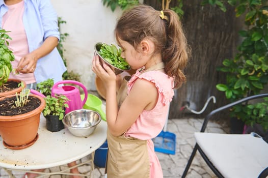 A young girl enjoys the fresh scent of herbs while gardening with an adult. The vibrant scene captures the joy and connection with nature in a homely garden setting.