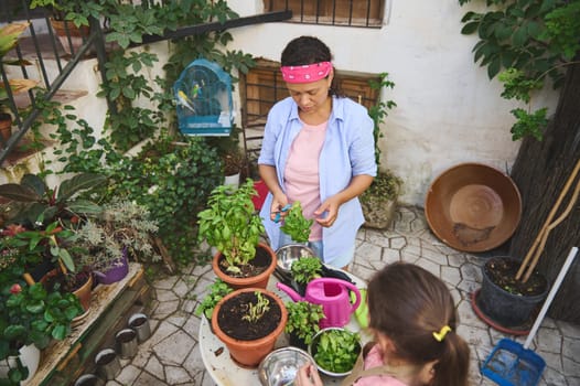 A mother and daughter engage in gardening, tending to herbs in a vibrant patio filled with potted plants. The scene exudes a sense of nature, family bonding, and tranquility.