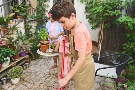 A child sweeping the floor while family members engage in gardening activities. Surrounded by lush plants and vibrant flowers, the setting is a cozy outdoor garden.