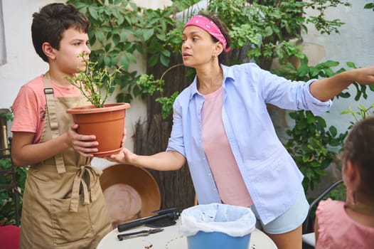 A mother guides her son in gardening, teaching him how to care for a potted plant. They bond while engaging in this outdoor activity, surrounded by greenery and sunlight.