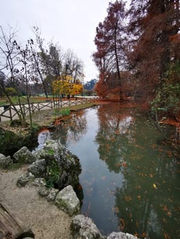 The scene features a serene park with a calm pond reflecting autumn leaves. Surrounding trees display vibrant fall colors, creating a peaceful atmosphere on a cloudy day.
