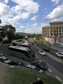 Numerous vehicles are caught in heavy traffic near the riverbank, with historic architecture in the background and a few scattered clouds overhead.