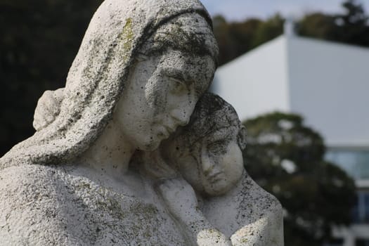 This artwork showcases a detailed sculpture of a mother and child, capturing a tender moment. The serene garden backdrop contrasts with the modern building behind, creating a unique atmosphere.