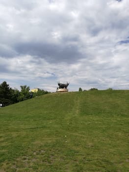 A prominent statue of an elk is situated at the top of a green hill, surrounded by a serene park. Visitors can be seen enjoying the lush landscape on a pleasant day.