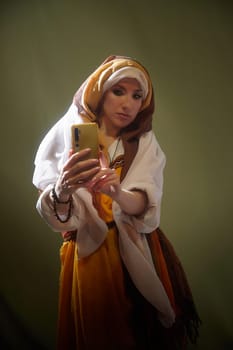 A young woman dressed in traditional eastern clothes poses thoughtfully, capturing herself on a smartphone during a creative shoot.