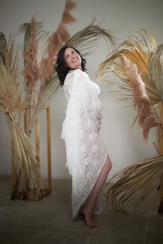 A cheerful woman stands confidently in an elegant dress against a backdrop of lush, dried plants, radiating a romantic vibe.