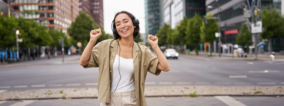 Dancing girl feeling happy in city. Asian woman dancing and listening music in headphones, posing on street.