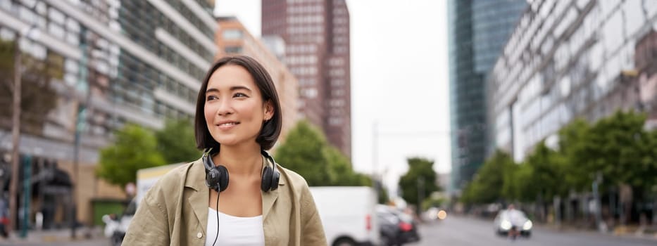 Portrait of young asian woman in headphones, posing in city, smiling and looking away, standing on street of city centre.