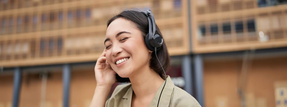 Portrait of beautiful asian woman in headphones, listening music on street of city centre, smiling happily.