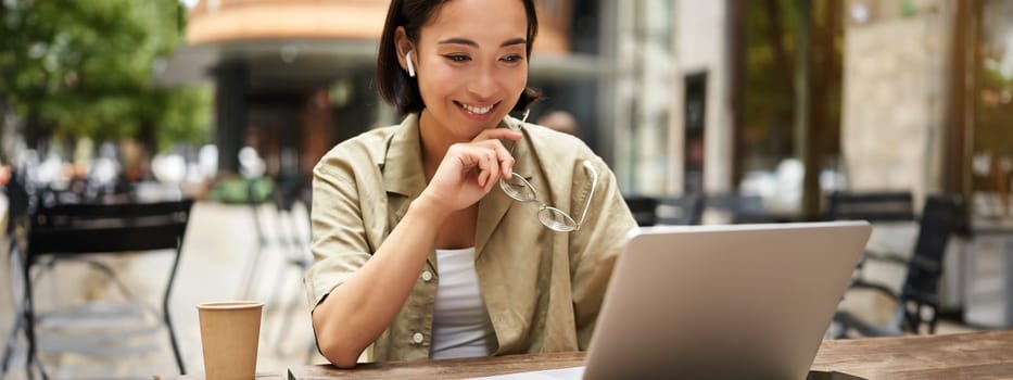 Smiling young korean woman looks at her laptop screen with pleased face, works remotely from outdoors, drinks coffee.