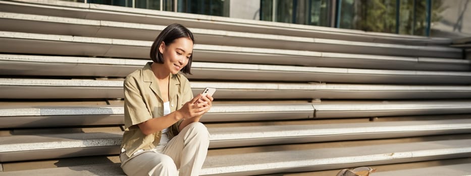 Beautiful asian girl sitting on stairs outside building, using mobile phone, looking at smartphone app and smiling.