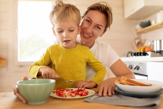 cheerful Caucasian mother and little son are sitting in the kitchen and preparing breakfast.