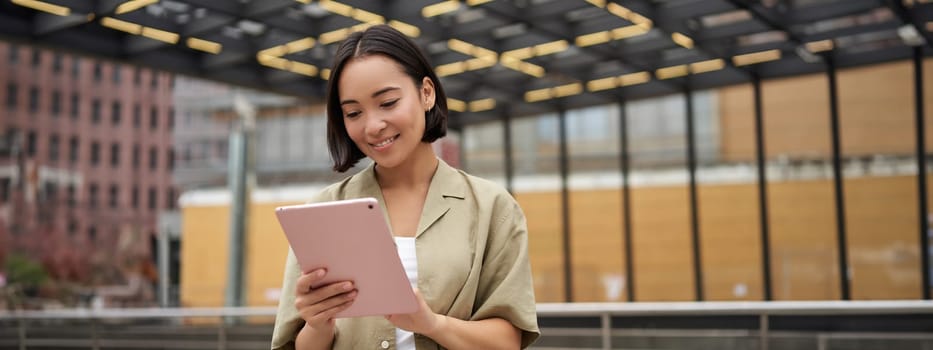 Portrait of beautiful asian girl using tablet, reading, looking at tablet while standing on street.