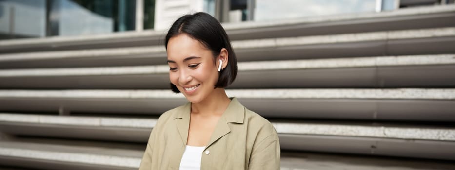 Remote worker. Smiling asian girl sits outdoors on street with laptop. Happy young woman working on computer remotely, listen music in wireless headphones.